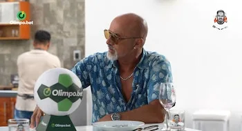 Carlos Alcántara viste una camisa azul y gafas de sol, sentado ante una mesa con un balón decorativo, copas y platos
