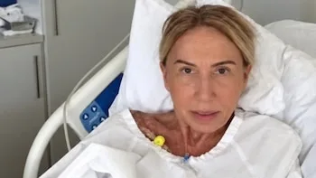Laura Bozzo recostada en una cama de hospital sobre almohadas blancas con bata médica y un catéter en el cuello